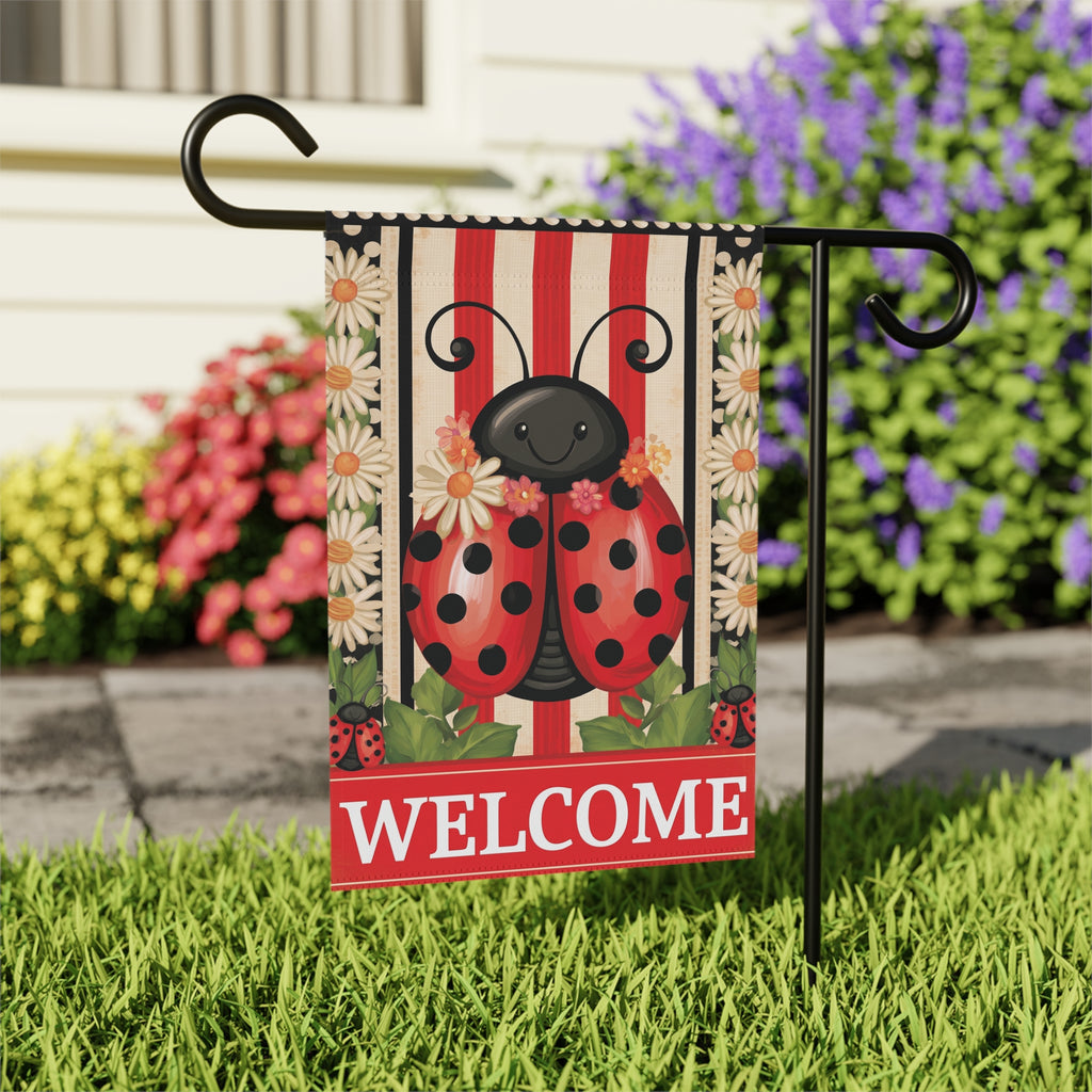 Close-up of the Welcome Ladybug garden flag on its stand near a stone walkway with purple flowering shrubs