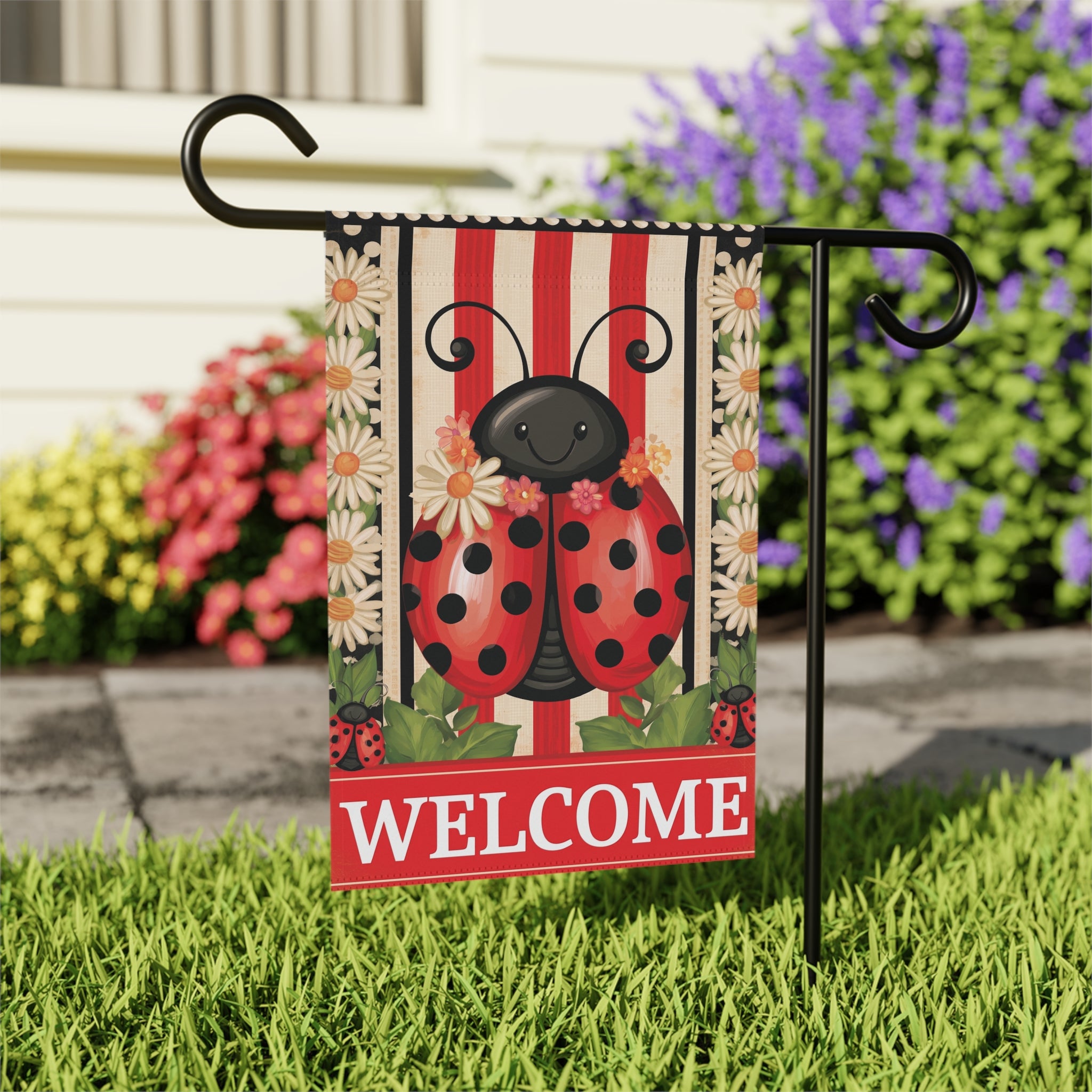 Close-up of the Welcome Ladybug garden flag on its stand near a stone walkway with purple flowering shrubs