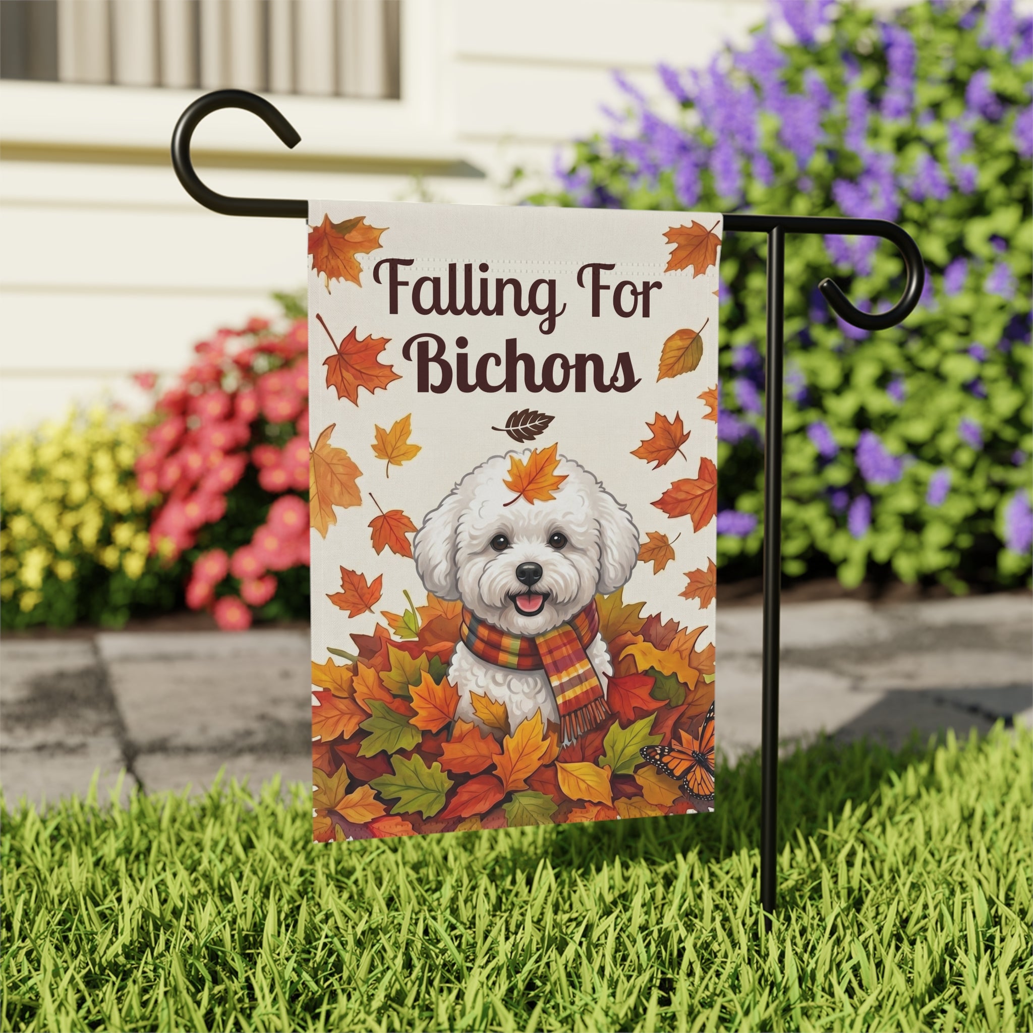 Close-up of the Falling For Bichons garden flag on its stand near a stone walkway with colorful flower bushes