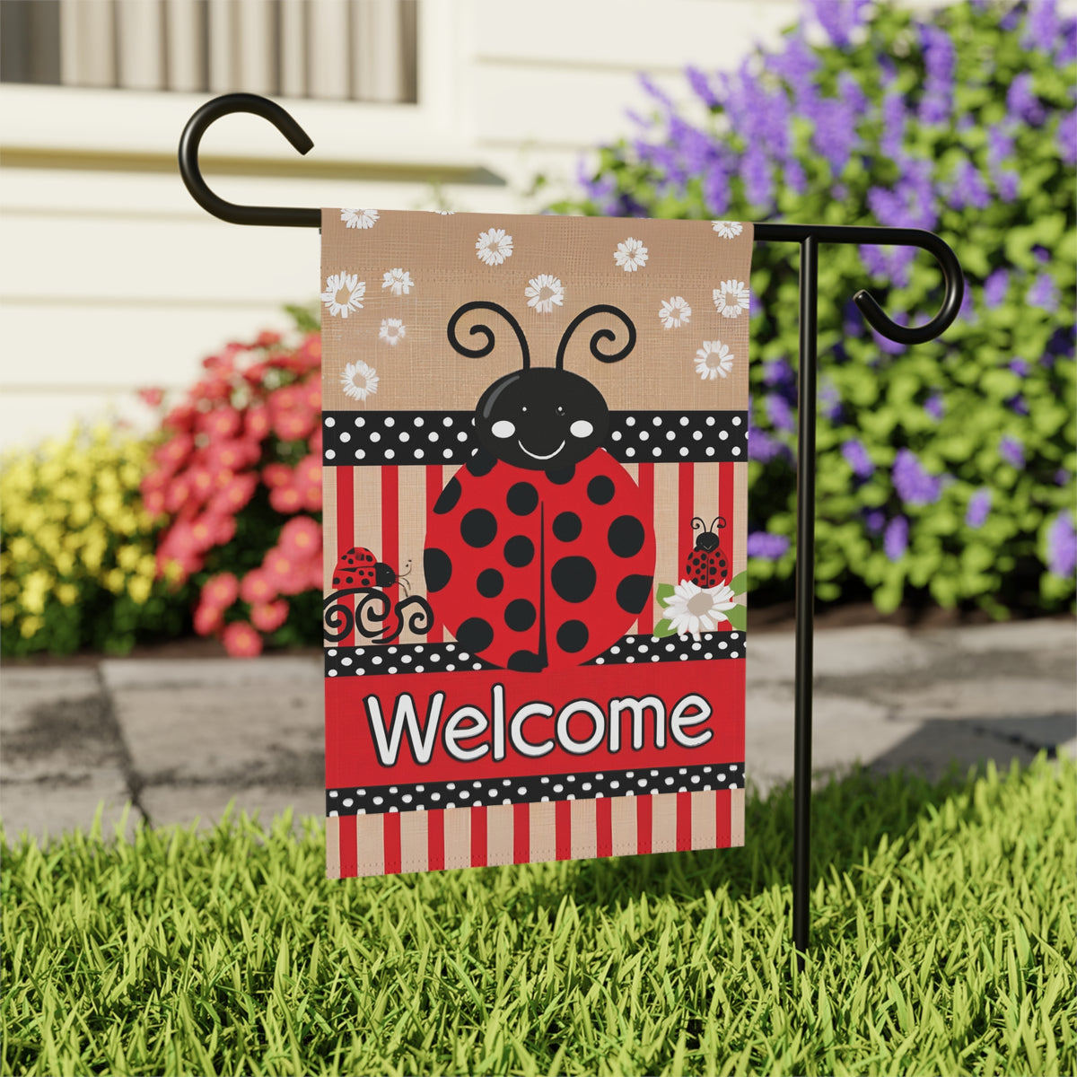 Close-up of the Large Ladybug Welcome garden flag on its stand beside a stone walkway and colorful flower beds
