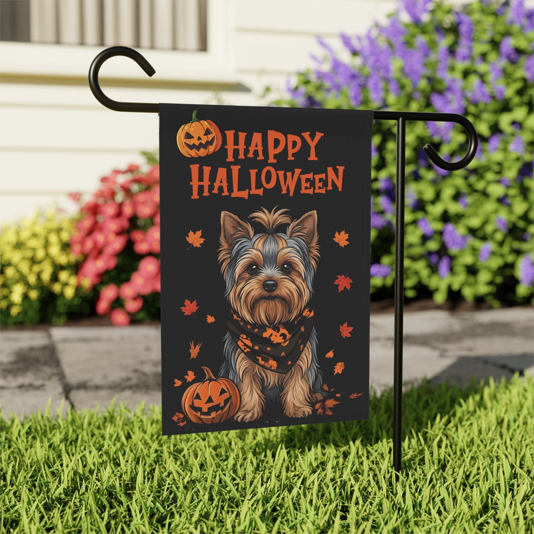 Close-up of the Little Pumpkin Yorkshire Terrier Halloween garden flag on its stand beside pink, yellow, and purple flowering shrubs near a walkway